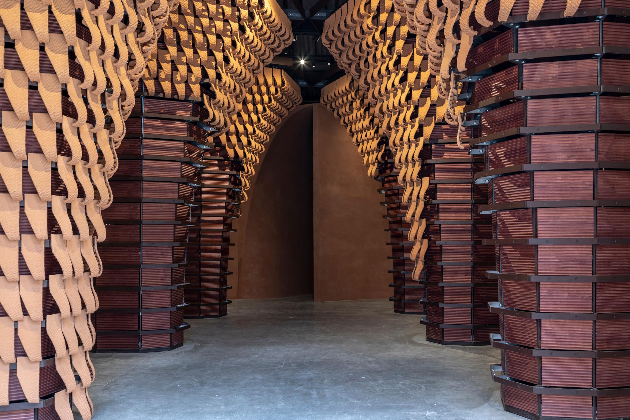 ARÁBIA SAUDITA - A mostra "IRTH" (legado), com a curadoria de Basma Bouzo e Noura Bouzo, explora a interação entre passado e futuro, a partir do elemento constitutivo da arquitetura saudita tradicional: a terra. 

A visita pelo pavilhão começa com o cruzamento de arcos metálicos, revestidos de madeira e ladrilhos, impressos em 3D em padrão irregular e ondulante, lembrando dunas do deserto. 

De um lado, os portais evocam arquiteturas monumentais, aparentemente indestrutíveis e duradouras; de outro, a transitoriedade da condição material, instável como a areia do deserto, apresentada na cobertura diminuindo gradualmente até desaparecer.
Expositor: Albara Osama Saimaldahar